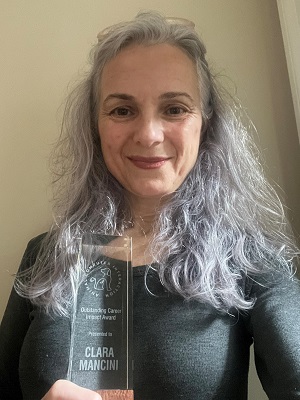 A woman with grey hair proudly holds up an award, smiling at the camera in a well-lit setting.