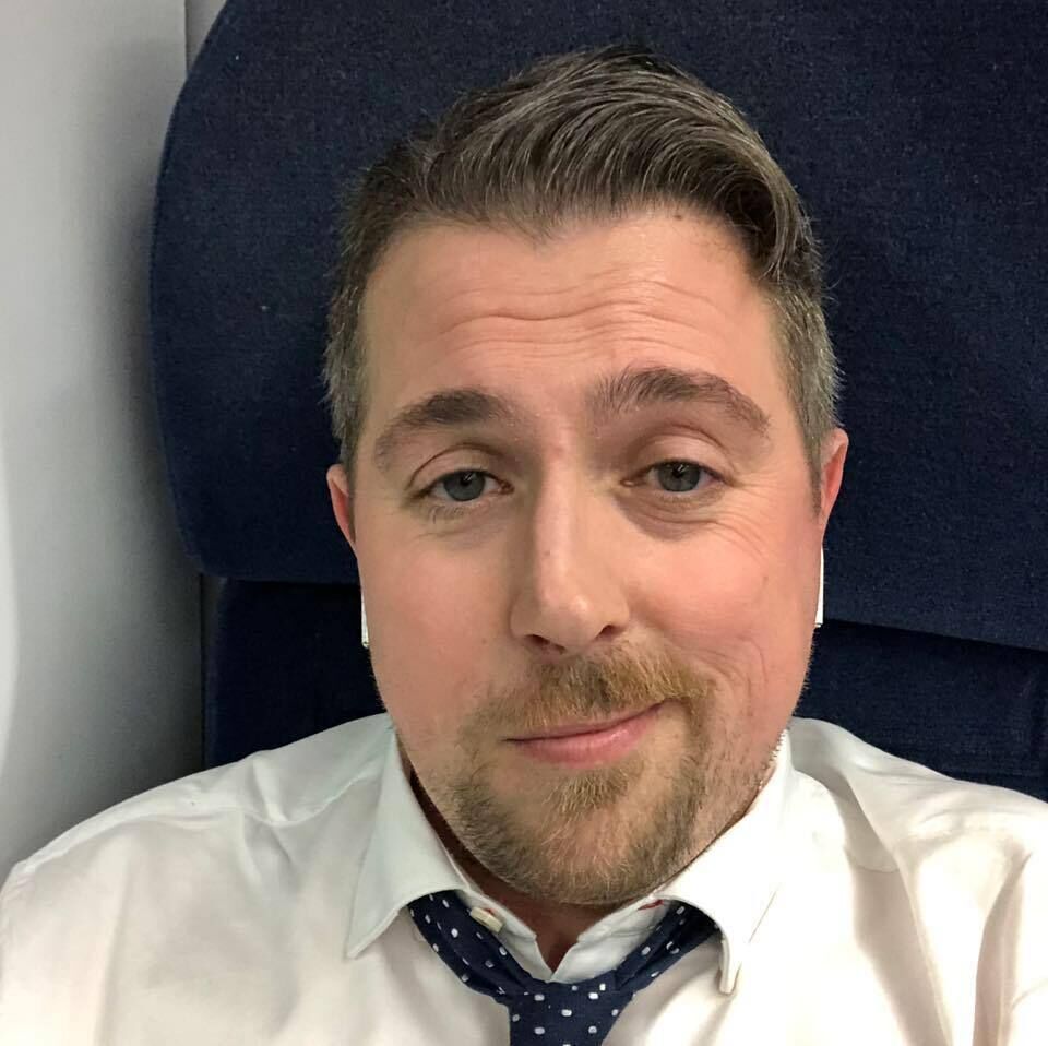 A man in a white shirt and tie seated on an airplane, looking out the window with a thoughtful expression
