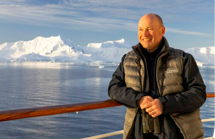 Feature image A man in a jacket and vest stands confidently on the deck of a ship, overlooking the water.