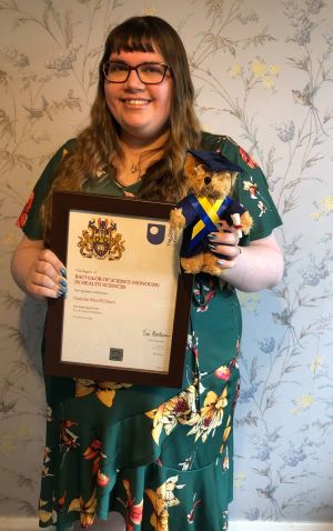 Feature image A woman proudly holds a certificate in one hand and a teddy bear in the other, smiling at the camera.
