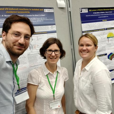 Three individuals stand in front of a colorful poster, engaged in conversation and looking at the displayed information.