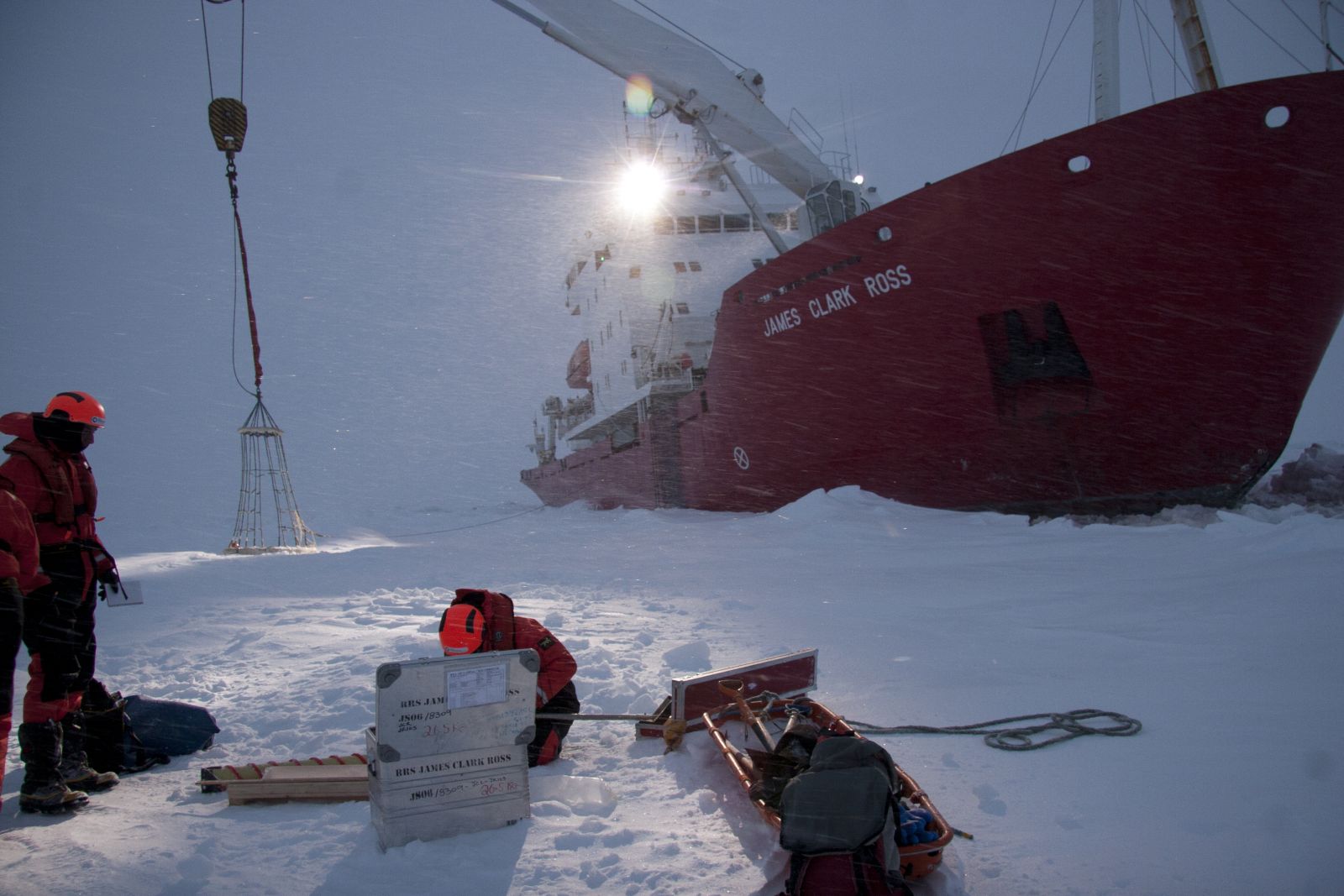 Researchers in red gear unload cargo via a net from the red icebreaker RRS James Clark Ross, moored in heavy snow and ice.
