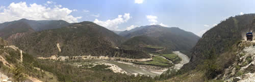 Panoramic view of a deep, forested mountain valley with a wide, winding river flowing through the center under a partly cloudy blue sky.