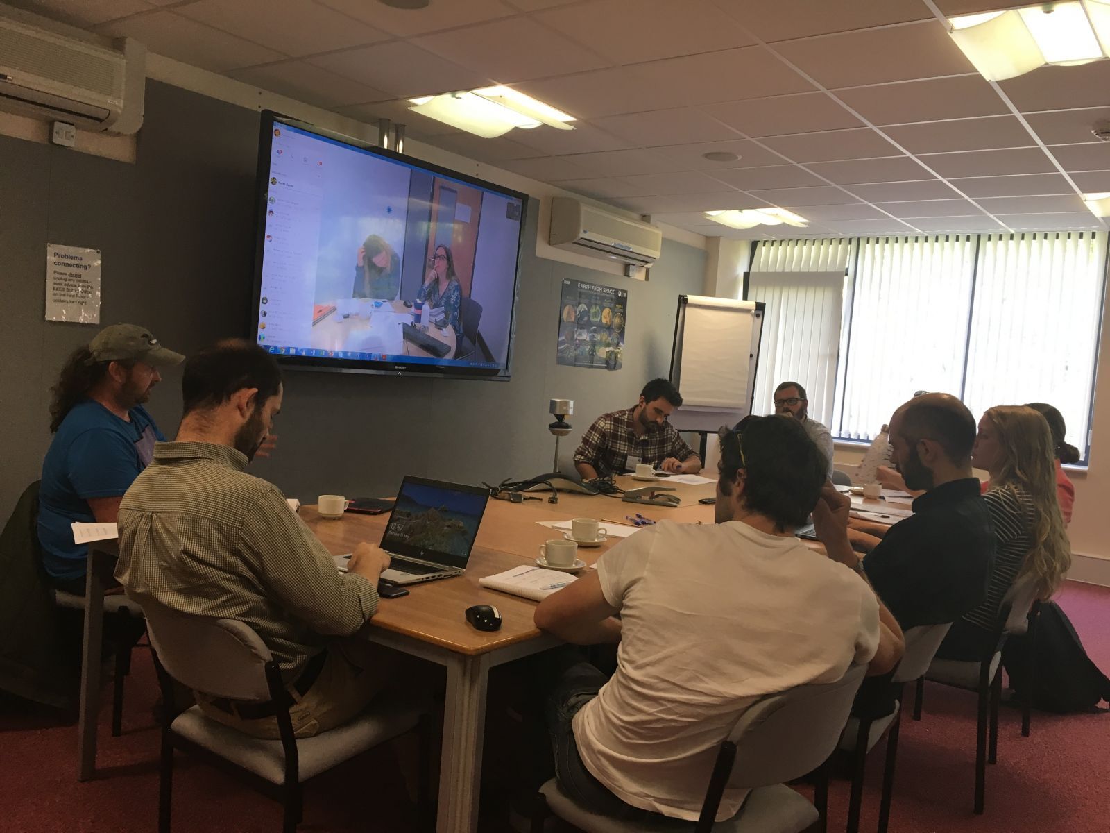 A group of people are gathered around a conference table participating in a video call displayed on a large wall-mounted television screen.