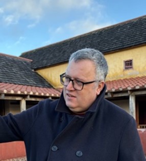 A man wearing glasses and a coat points to an object, indicating interest or directing attention to it.