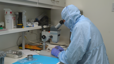 Technician in full light-blue cleanroom suit examining a sample under a microscope in a laboratory setting.