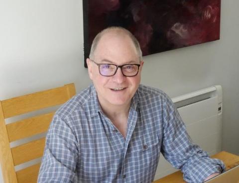 Smiling, middle-aged man with glasses wearing a blue plaid shirt sits indoors next to a wooden chair and a white wall heater, with abstract art above.