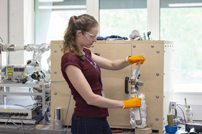 A woman wearing orange gloves operates a machine, focused on her task in a workshop environment.