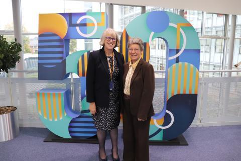 Two women stand together in front of a large number sign, smiling and engaging with each other.