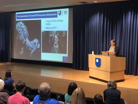 A woman stands at a podium, presenting to an audience at a conference, with slides displayed in the background.