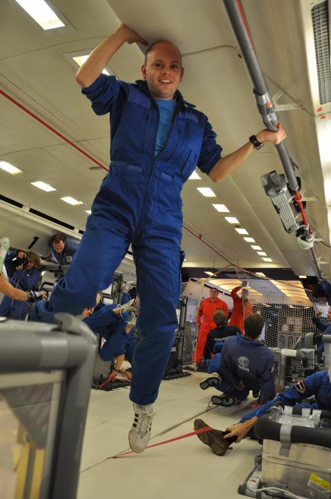 A man in blue overalls hangs from the ceiling of an airplane, performing maintenance work in a confined space.